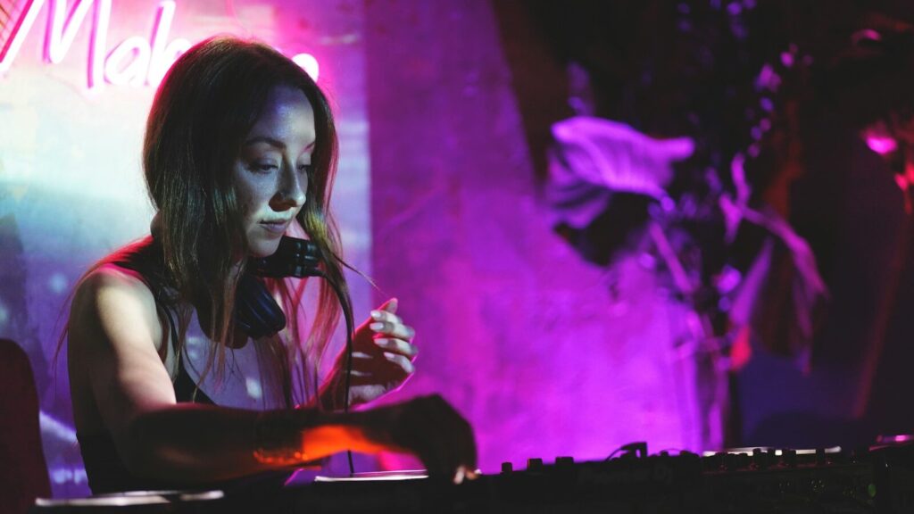 a woman sitting at a keyboard in front of a neon sign What is DJing